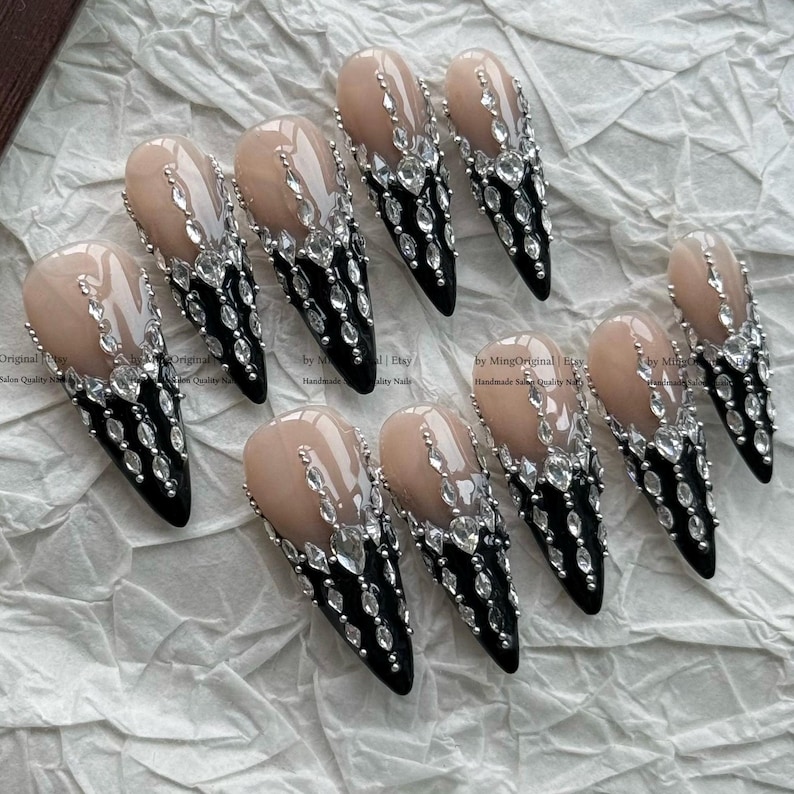 May include: A set of ten stiletto-shaped press-on nails featuring a nude base transitioning into a black tip. The nails are embellished with clear, faceted rhinestones arranged in a vertical pattern that extends from the nude area down to the black tips.