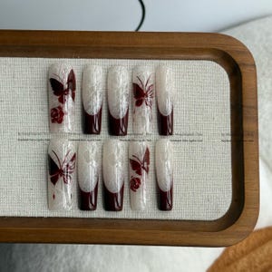 Pu&ograve; includere: Un set di dieci unghie finte con un design in pizzo bianco, punte bordeaux e dettagli di farfalle e rose. Le unghie sono disposte su un vassoio di legno con uno sfondo in tessuto neutro. Handmade salon quality nails.