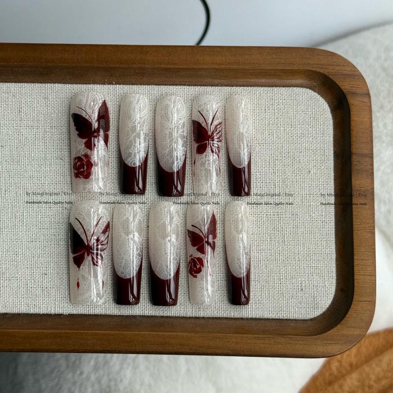 Pu&ograve; includere: Un set di dieci unghie finte con un design in pizzo bianco, punte bordeaux e dettagli di farfalle e rose. Le unghie sono disposte su un vassoio di legno con uno sfondo in tessuto neutro. Handmade salon quality nails.