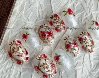 May include: Oval-shaped artificial nails with a white pearlescent base, adorned with hand-painted red roses and gold-colored decorative frames. The text "Ming Original" is visible on some nails. A floral and ornate design.