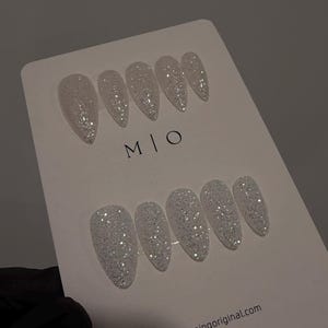 May include: A set of twelve white glitter press-on nails in various sizes, arranged on a white card. The nails have a glossy finish and are shaped in a pointed oval. The card has the letters "M|O" printed on it.