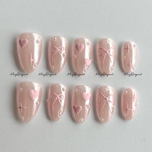Könnte beinhalten: Ein Set aus zehn künstlichen Nägeln in einem hellrosa Farbton, die jeweils mit filigranen Designs verziert sind. Die Nägel weisen geprägte Herzen, Schleifen und Sternmuster auf, die mit kleinen perlenartigen Verzierungen akzentuiert sind. Der Text "Ming Original" ist auf einigen Nägeln sichtbar.