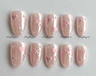 May include: A set of ten artificial nails in a light pink hue, each adorned with delicate designs. The nails feature embossed hearts, bows, and starburst patterns, accented with small pearl-like embellishments. The text "Ming Original" is visible on some nails.