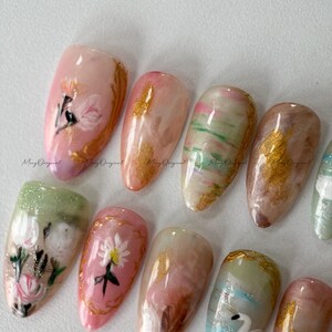 May include: A collection of almond-shaped artificial nails with various designs. Some nails feature floral art in pink, white, and gold. Others have marble effects in shades of pink, green, and brown. The text "Ming Original" is visible on some nails.