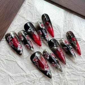 May include: A set of ten long, almond-shaped artificial nails. Each nail features a design with a gradient from black to red, adorned with rose patterns, silver accents, and pearl embellishments. The nails are arranged on a light-colored surface.