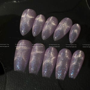 May include: A set of ten lavender-colored, oval-shaped false nails. Each nail has a shimmering, glittery effect with streaks of silver and blue. The nails are arranged on a dark surface. Text on the image reads "MingOriginal | Etsy Handmade Salon Quality Nails."