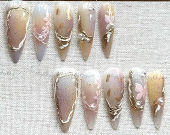 May include: A collection of ten long, almond-shaped artificial nails. Each nail features a gradient design with shades of beige, white, and gold. Some nails have floral and pearl accents, while others have gold foil and glitter.