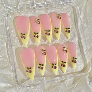 May include: Set of ten artificial nails with a pink and yellow design. Each nail features a pink tip, a yellow base, and a cherry charm with green leaves and purple gems. The nails are displayed in a clear plastic case.