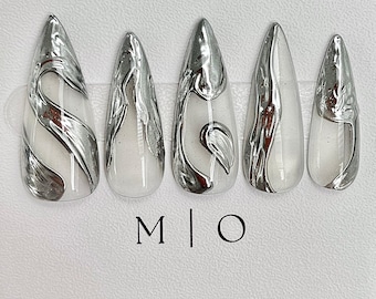 May include: Five silver and white nail tips with a marbled design. The tips are arranged in a row on a white surface. The text "M | O" is printed below the tips.