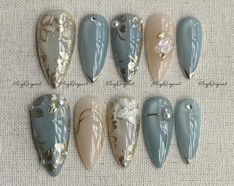 May include: A set of ten artificial nails in various designs. The nails feature a combination of light blue, beige, and gold accents. Some nails have floral patterns, while others have gold lines or small embellishments. The text "Ming Original" is printed on the nails.