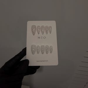 K&ouml;nnte beinhalten: Ein Set silberner Glitzer-Press-On-N&auml;gel auf einer wei&szlig;en Karte. Die Karte tr&auml;gt den Text "M | O" und die Website-Adresse "www.mingoriginal.com". Die N&auml;gel sind in zwei Reihen zu je sechs St&uuml;ck angeordnet und haben eine spitze Form.