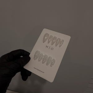 K&ouml;nnte beinhalten: Auf einer wei&szlig;en Karte werden zwei Sets silberner Glitzer-Press-On-N&auml;gel in Mandel- und Ovalform pr&auml;sentiert. Die Karte tr&auml;gt den Text "M | O" und die Website-Adresse "www.mingoriginal.com".