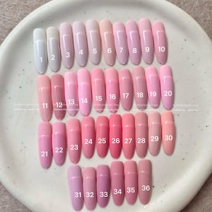 May include: A collection of 36 artificial nails in various shades of pink, arranged on a white ceramic plate. Each nail is numbered, ranging from light pink to deeper rose hues. The nails are oval-shaped and have a glossy finish.