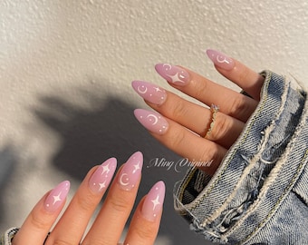 May include: Close-up of a hand with light pink almond-shaped nails with white crescent moon and star designs. The hand is wearing a silver ring with a white stone.