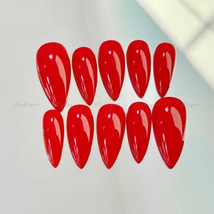 May include: A set of ten glossy, bright red, almond-shaped artificial nails. The nails are arranged in two rows, with a slight curve and a pointed tip. The nails are evenly spaced and reflect light.