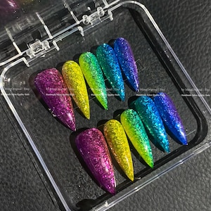 Peut inclure: Un ensemble de dix faux ongles longs recouverts de paillettes dans un étui en plastique transparent. Les ongles sont disposés en deux rangées de cinq, présentant un arc-en-ciel de couleurs : violet, jaune, vert, bleu et un mélange.