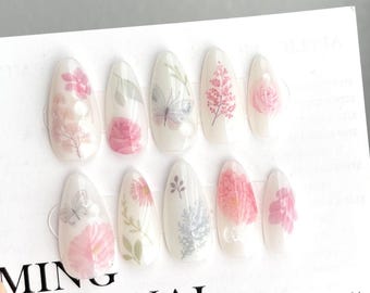 May include: A set of ten white oval-shaped fake nails with floral and butterfly designs. The nails are arranged in two rows on a white background. The text "MING ORIGINAL" and "Make refinement a habit" are printed on the background.