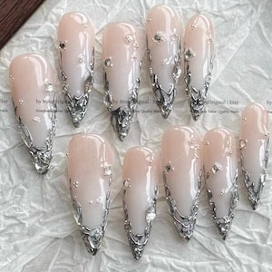 May include: A set of ten almond-shaped press-on nails featuring a pale pink to white gradient base. Each nail is adorned with metallic silver liquid-style accents and small clear rhinestones. The text 'by MingOriginal | Etsy Handmade Salon Quality Nails' is visible.