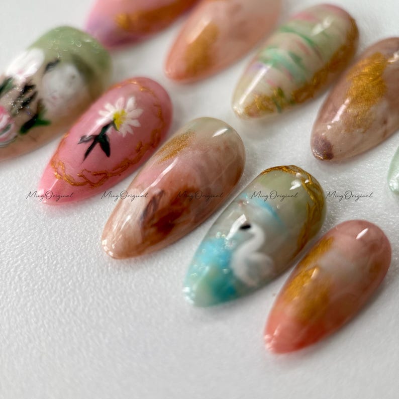 May include: A collection of artificial nails with various designs. Some nails feature floral patterns, others have abstract swirls, and some showcase gold accents. The nails are in shades of pink, peach, green, and gold. The text "Ming Original" is printed on some nails.
