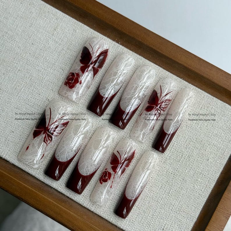 Pu&ograve; includere: Un set di dieci unghie finte con un design in pizzo bianco e dettagli bordeaux. Ogni unghia presenta un disegno a farfalla o rosa in bordeaux. Le unghie sono esposte su un vassoio di legno.