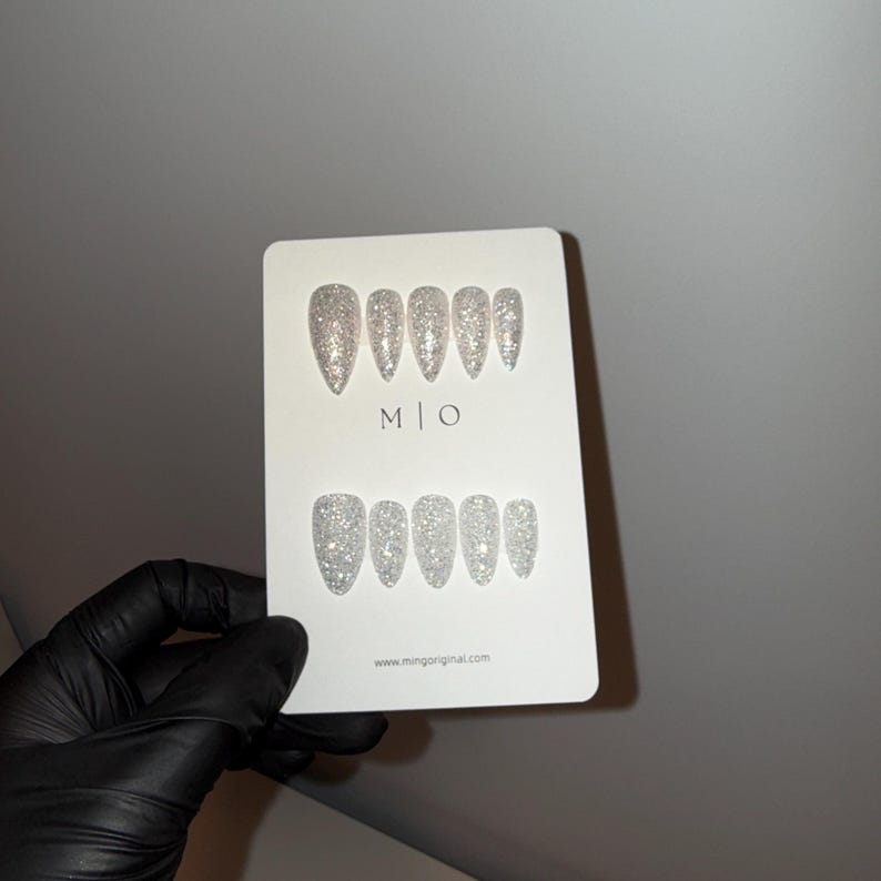 K&ouml;nnte beinhalten: Ein Set silberner Glitzer-Press-On-N&auml;gel in verschiedenen Gr&ouml;&szlig;en, pr&auml;sentiert auf einer wei&szlig;en Karte. Die Karte tr&auml;gt den Text "M | O" und die Website "www.mingoriginal.com". Die N&auml;gel sind mandelf&ouml;rmig und haben ein funkelndes, schillerndes Finish.