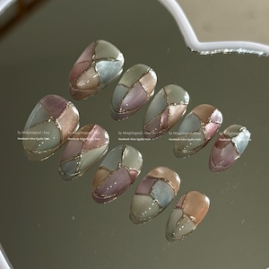 Op de afbeelding: Een set van tien ovale kunstnagels. Elke nagel heeft een mozaïekontwerp met secties in gedempt roze, beige en blauwgrijs, omlijnd met goud. De nagels zijn gerangschikt op een reflecterend oppervlak.