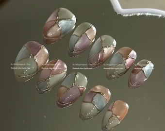 May include: A set of ten oval-shaped, press-on nails. Each nail features a mosaic design with sections of muted pink, beige, and blue-grey, outlined with gold. The nails are arranged on a reflective surface.