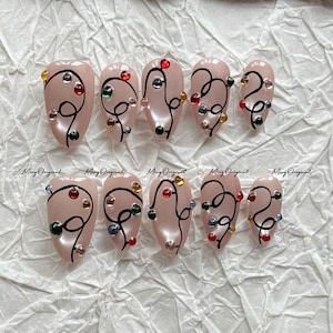 Peut inclure: Dix faux ongles avec une finition nacrée rose clair. Chaque ongle présente un motif de lignes noires avec des strass colorés. Le texte "Ming Original" est imprimé au bas de chaque ongle.