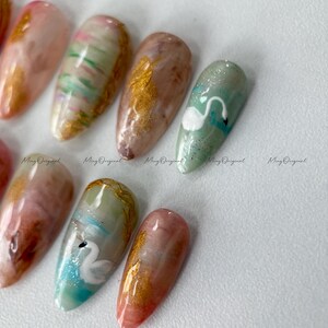 May include: A collection of almond-shaped artificial nails with various designs. Some nails feature gold foil accents, while others showcase abstract patterns in shades of pink, green, and brown. One nail has a white swan design.