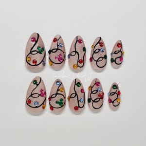 May include: Set of ten almond-shaped press-on nails. Each nail is a glittery nude color, adorned with a black swirled design and colorful rhinestones. The nails are arranged on a white surface.