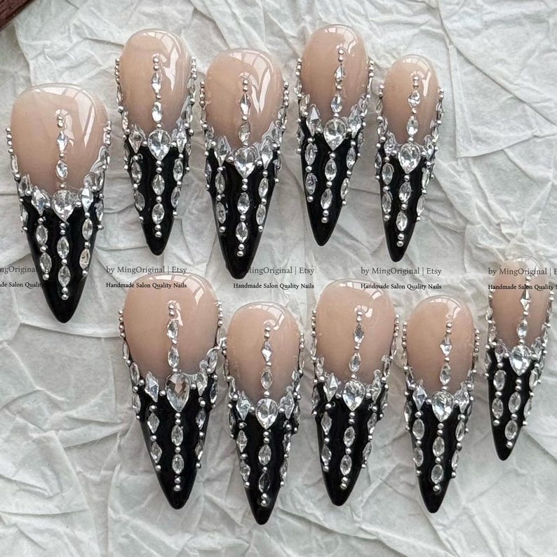 May include: A set of ten stiletto-shaped press-on nails featuring a nude base and a black tip. Each nail is decorated with clear, faceted rhinestones arranged in a vertical, cascading pattern that extends from the nude area down to the black tip.