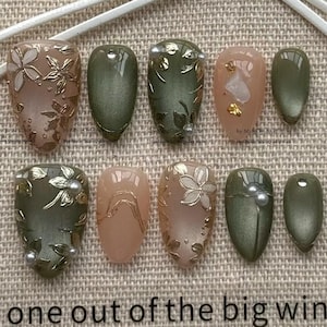 May include: A set of ten press-on nails featuring a color palette of olive green and soft nude pink. The designs include metallic gold floral embossed patterns, small white pearl accents, gold leaf flakes, and solid olive green finishes.
