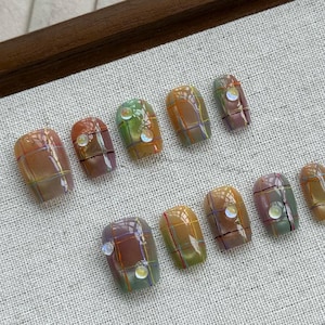 Op de afbeelding: Tien vierkante kunstnagels met een ruitpatroon in tinten bruin, oranje, groen en blauw. Elke nagel heeft een klein, iriserend steentje in het midden.