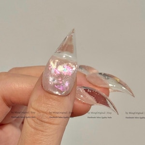 Può includere: Primo piano di unghie acriliche trasparenti e appuntite. Un'unghia presenta una base rosa chiaro con glitter iridescenti. Le altre unghie sono trasparenti e appuntite. Unghie di qualità da salone fatte a mano.
