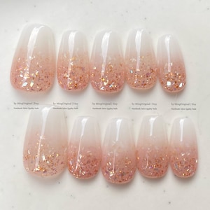 Puede incluir: Diez uñas postizas con un degradado de blanco a rosa y purpurina dorada. Las uñas están moldeadas y diseñadas para ser aplicadas. El texto "by MingOriginal | Etsy Handmade Salon Quality Nails" es visible.