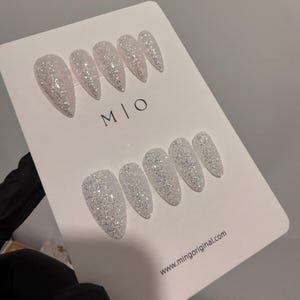 K&ouml;nnte beinhalten: Ein Set aus silbernen Glitzer-Press-On-N&auml;geln in Stiletto-Form. Die N&auml;gel sind auf einer wei&szlig;en Karte mit dem Text "M | O" und "www.mingoriginal.com" angeordnet. Die N&auml;gel sind hellsilberfarben und haben ein funkelndes Finish.