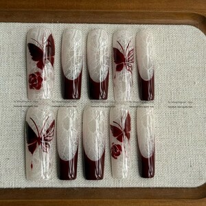 Pu&ograve; includere: Un set di dieci unghie finte con un design in pizzo bianco, punte bordeaux e accenti di farfalle e rose bordeaux. Le unghie sono rettangolari e hanno una finitura lucida.