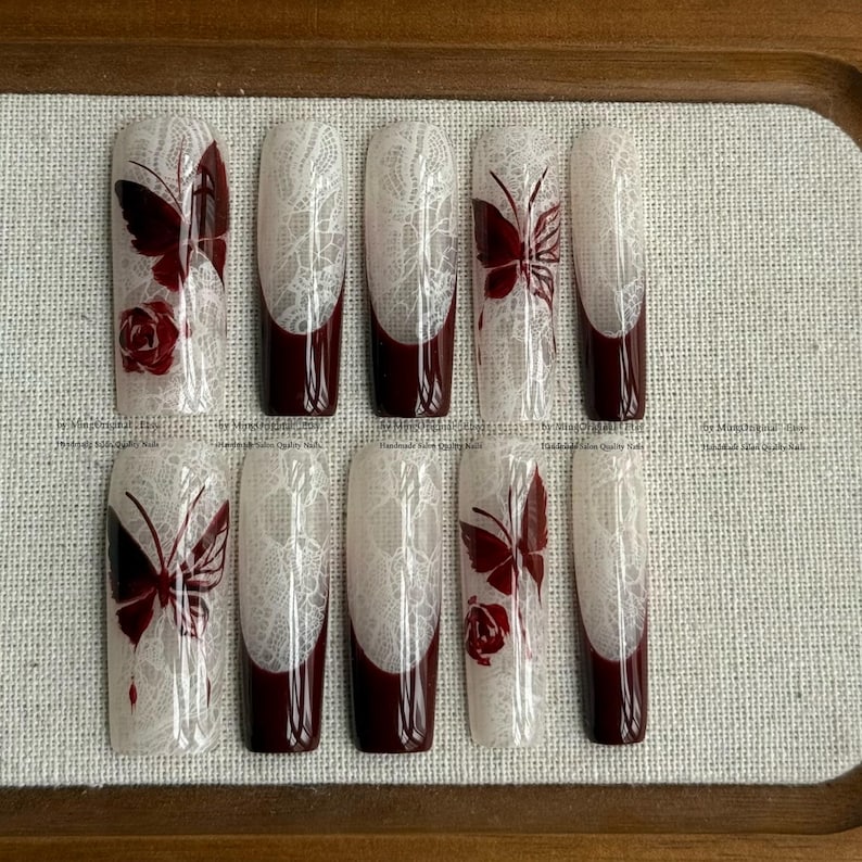 Pu&ograve; includere: Un set di dieci unghie finte con un design in pizzo bianco, punte bordeaux e accenti di farfalle e rose bordeaux. Le unghie sono rettangolari e hanno una finitura lucida.
