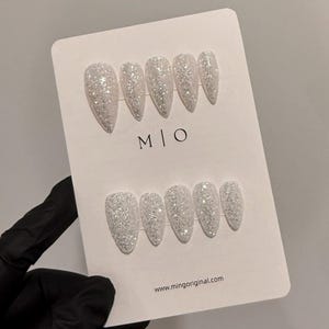 K&ouml;nnte beinhalten: Ein Set glitzernder, wei&szlig;er Kunstn&auml;gel in Stilettoform, angeordnet auf einer wei&szlig;en Karte. Die Karte zeigt die Buchstaben "M | O" und die Website-Adresse "www.mingoriginal.com". Die N&auml;gel werden von einer behandschuhten Hand gehalten.