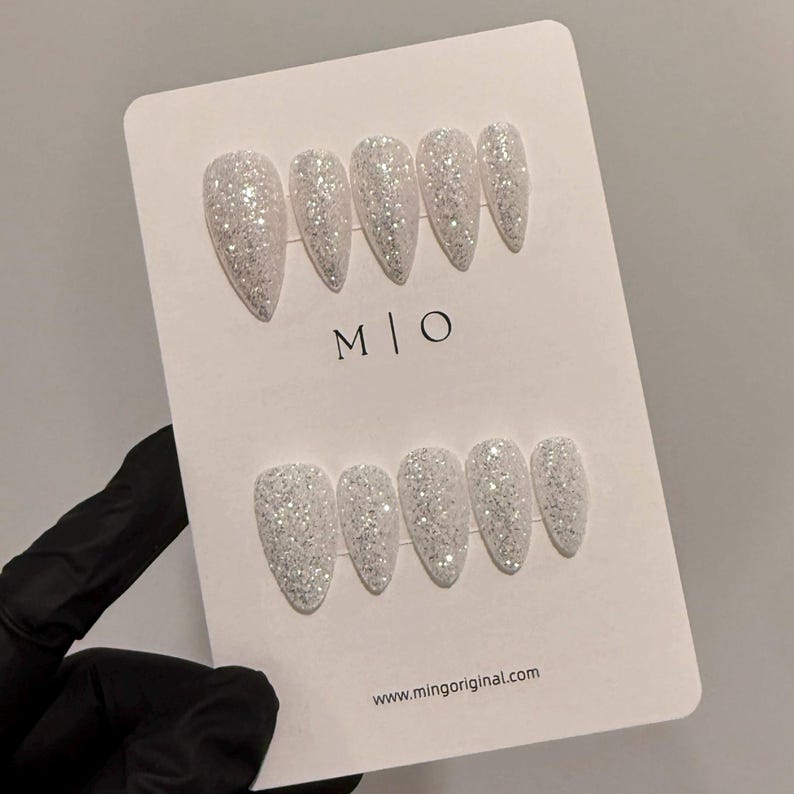 K&ouml;nnte beinhalten: Ein Set glitzernder, wei&szlig;er Kunstn&auml;gel in Stilettoform, angeordnet auf einer wei&szlig;en Karte. Die Karte zeigt die Buchstaben "M | O" und die Website-Adresse "www.mingoriginal.com". Die N&auml;gel werden von einer behandschuhten Hand gehalten.