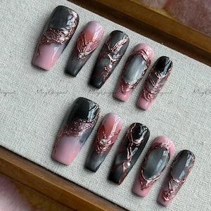 May include: A set of ten long, coffin-shaped artificial nails. Each nail features a gradient design in shades of pink, gray, and black, accented with rose gold filigree and small embellishments. The nails are arranged on a light-colored surface.
