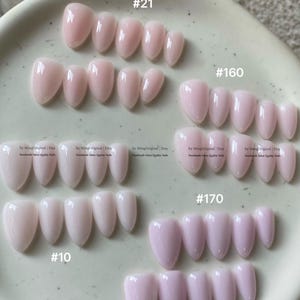 以下が含まれることがあります： 斑点のある表面にグループ分けされた、光沢のある淡いピンク色のアーモンド型プレスオンネイルのセット。グループには#21、#160、#170、#10のラベルが付いています。各セットにはさまざまなサイズが含まれており、一部にはby MingOriginal | Etsyというテキストが記載されています。