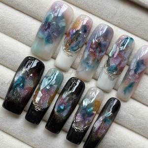 May include: Ten long, artificial nails with a glossy finish. The nails display abstract designs in blue, purple, and gold, some with silver embellishments. The nails are arranged on a light-coloured surface. The designs are reminiscent of a galaxy.