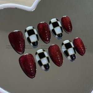 May include: Set of ten artificial nails in a variety of designs. Some nails are a deep red with glitter, while others feature a black and white checkerboard pattern with gold accents. The nails are oval-shaped.
