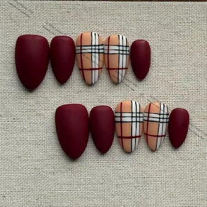 May include: A set of matte burgundy and plaid design press-on nails. The plaid nails feature a beige base with black, white, and red lines. The nails are oval-shaped and arranged on a neutral fabric surface.