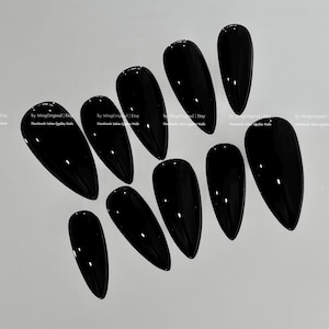May include: A set of ten glossy, black, almond-shaped press-on nails. The nails are arranged on a light gray surface, showcasing their smooth, reflective finish. The nails are a solid, opaque black color.