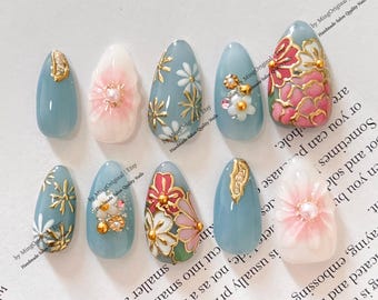 May include: A collection of ten artificial nails in various designs. The nails feature a light blue, pink, and white color palette with gold and white floral and abstract accents. Some nails have 3D floral embellishments.
