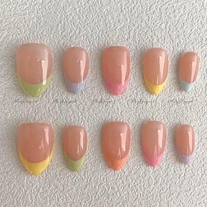 Peut inclure: Un ensemble de dix faux ongles de différentes tailles, chacun avec une base nude et un bout français coloré. Les bouts sont peints en vert, violet, rose, jaune et bleu. Les ongles sont disposés sur une surface blanche texturée.
