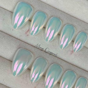 May include: Ten iridescent white oval-shaped fake nails with a pearlescent finish. The nails are arranged in two rows on a white surface. The text "Mug Original" is visible in the image.