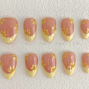 Peut inclure: Dix faux ongles ovales avec une base rose clair, des pointes jaunes et des motifs floraux jaunes. Chaque ongle est orné d'une petite perle dorée. Finition brillante.
